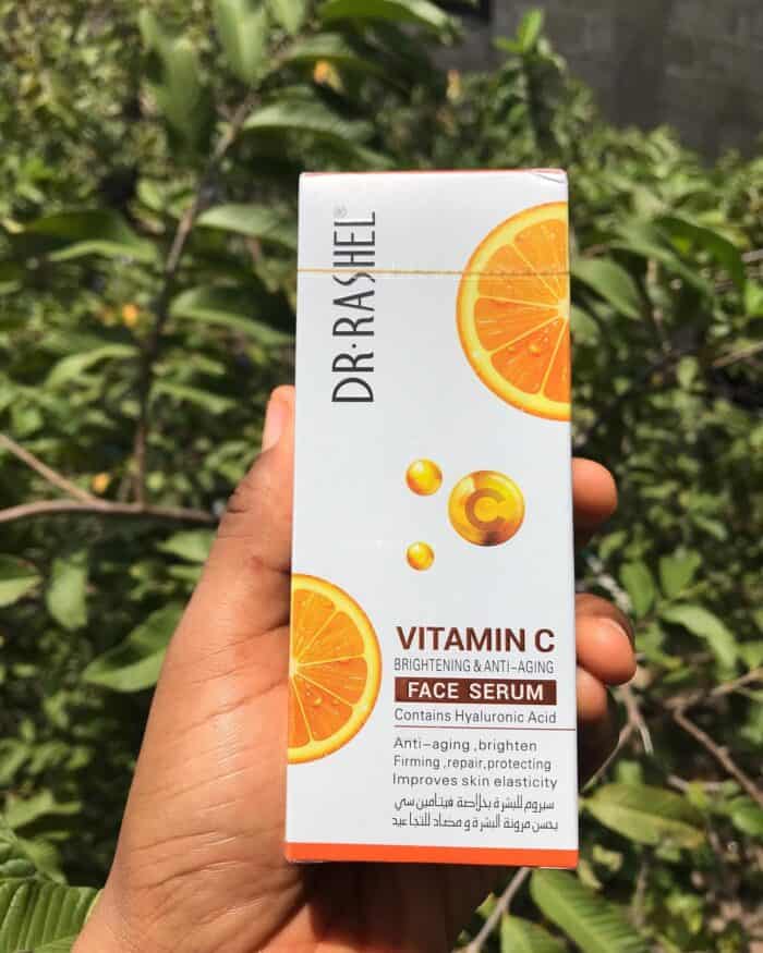 Person holding Dr Rashel Vitamin C Face Serum box with orange slice design, showing brightening and anti-aging skincare benefits