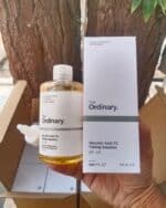 Hand holding The Ordinary Glycolic Acid 7% Toning Solution bottle next to its white packaging box, both displayed outdoors with tree bark in the background.