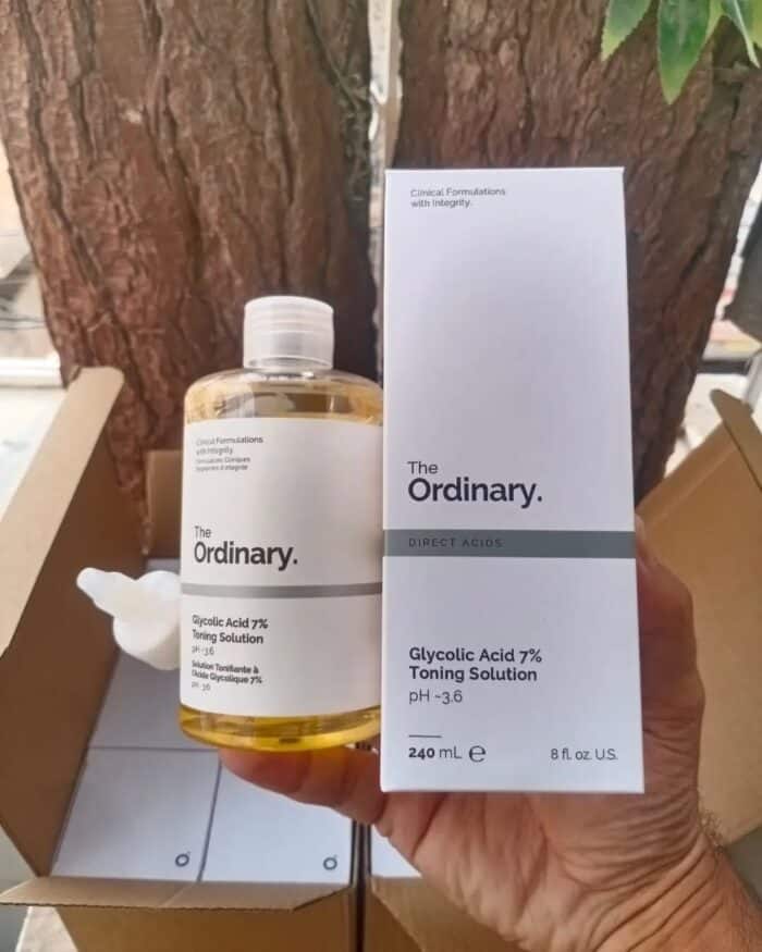Hand holding The Ordinary Glycolic Acid 7% Toning Solution bottle next to its white packaging box, both displayed outdoors with tree bark in the background.