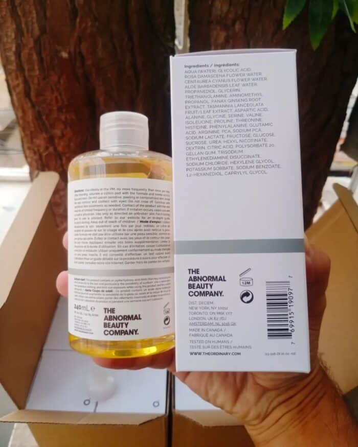 Hand holding The Ordinary Glycolic Acid 7% Toning Solution 240ml bottle next to its box, both positioned vertically outdoors with tree background