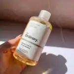 Hand holding a bottle of The Ordinary Glycolic Acid 7% Toning Solution with a clear label, partially filled with yellow-tinted liquid, against a softly lit background