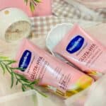 Two pink tubes of Vaseline Gluta-Hya Dewy Radiance Lotion 330ml placed on soft fabric with a small clock and green leaves in the background
