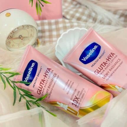 Two pink tubes of Vaseline Gluta-Hya Dewy Radiance Lotion 330ml placed on soft fabric with a small clock and green leaves in the background