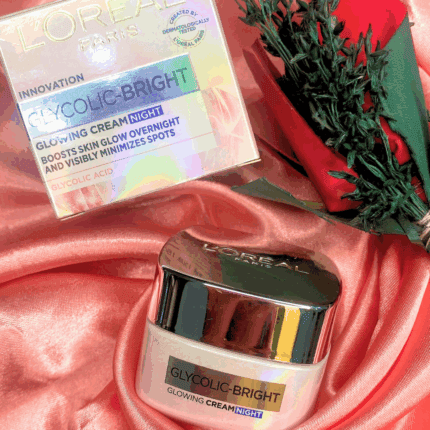 L’Oréal Glycolic-Bright Glowing Night Cream jar with silver lid placed beside its holographic box packaging, displayed on pink satin fabric with a red rose for an elegant skincare presentation.