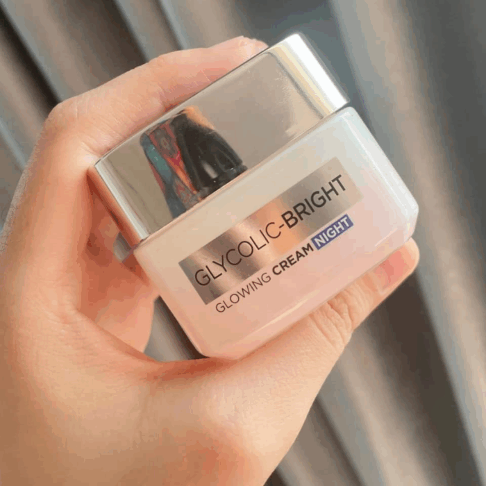 A hand holding the L’Oréal Glycolic-Bright Glowing Night Cream with a shiny silver lid and square jar, shown against soft curtain background in natural lighting.