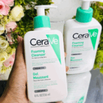 Hand holding CeraVe Foaming Cleanser 236ml for normal to oily skin, fragrance-free face wash with ceramides, niacinamide, and hyaluronic acid, second bottle in background with floral decoration.