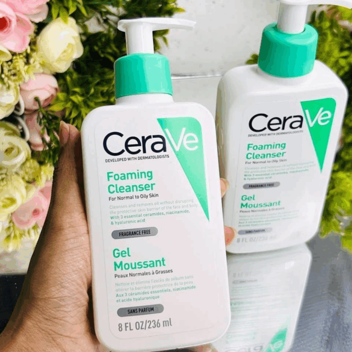 Hand holding CeraVe Foaming Cleanser 236ml for normal to oily skin, fragrance-free face wash with ceramides, niacinamide, and hyaluronic acid, second bottle in background with floral decoration.