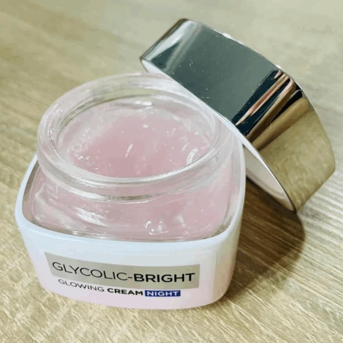 An open jar of L’Oréal Glycolic-Bright Glowing Night Cream showing its pink gel-cream texture, with the silver lid placed on the side on a wooden surface.