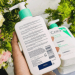 Back view of CeraVe Foaming Cleanser 236ml bottle showing ingredients list, barcode, and usage directions, held in hand with floral background and another cleanser bottle beside it.