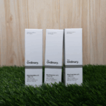 Three boxes of The Ordinary Niacinamide 10% + Zinc 1% serum 30ml displayed upright on green grass with a wooden background, showing high-strength vitamin and mineral blemish control formula packaging.