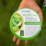 A hand holding Dr Rashel Aloe Vera Soothing & Moisturizing Gel tub with green lid, aloe leaf graphics, and listed benefits such as hydration, glowing skin, cooling effect, and moisturizing care. Green grass background.