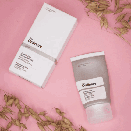 The Ordinary Azelaic Acid Suspension 10% shown with grey squeeze tube and minimalist white box packaging, placed on a pink background with dried oat stems for a natural aesthetic.