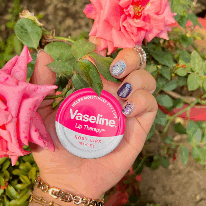 A hand with colorful glitter nail art and a gold bracelet holding Vaseline Lip Therapy Rosy Lips balm tin (pink and white) next to blooming pink roses and green leaves in a garden setting.