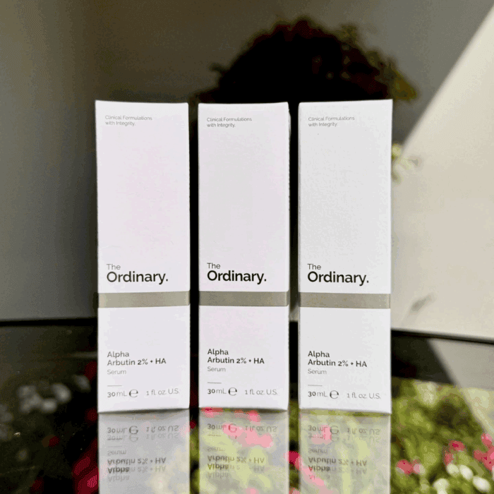 Three boxes of The Ordinary Alpha Arbutin 2% + HA Serum (30ml each) displayed upright on a reflective glass surface, with a blurred natural background.