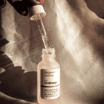 The Ordinary Alpha Arbutin 2% + HA Serum in a frosted glass bottle with an open dropper showing clear serum, placed against a textured beige satin background with soft shadows.