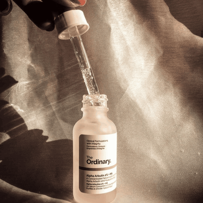 The Ordinary Alpha Arbutin 2% + HA Serum in a frosted glass bottle with an open dropper showing clear serum, placed against a textured beige satin background with soft shadows.