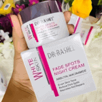 A hand holding Dr Rashel White Skin Fade Spots Night Cream jar and its matching box, featuring Arbutin and Niacinamide for even skin tone, dark spot reduction, and nourishing repair. Color scheme white, pink, and silver with floral background.
