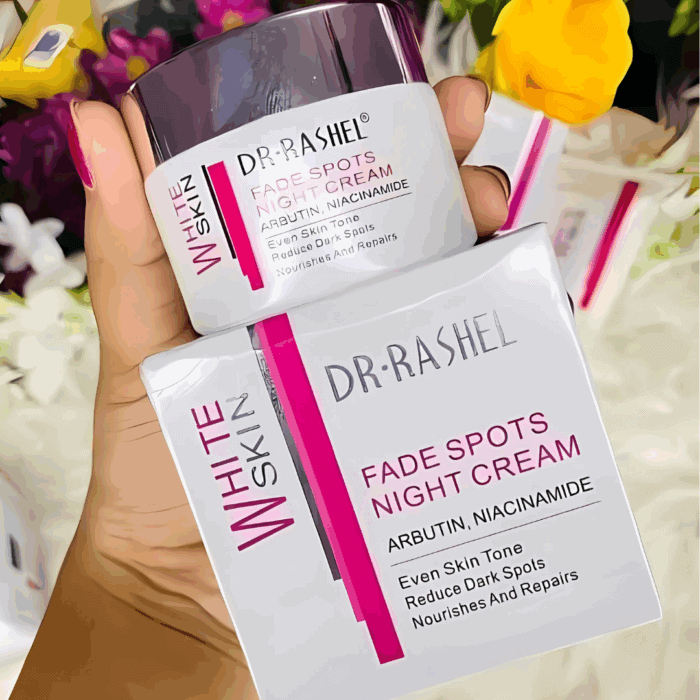 A hand holding Dr Rashel White Skin Fade Spots Night Cream jar and its matching box, featuring Arbutin and Niacinamide for even skin tone, dark spot reduction, and nourishing repair. Color scheme white, pink, and silver with floral background.
