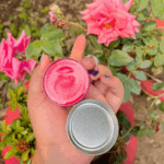 A hand with purple glitter nail art holding an open tin of Vaseline Lip Therapy Rosy Lips balm, showing the smooth pink product inside, with blooming pink roses and green leaves in the background.