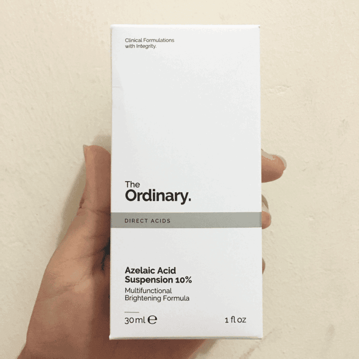 Hand holding The Ordinary Azelaic Acid Suspension 10% box packaging (30ml) with minimalist white design, labeled as a multifunctional brightening formula, photographed against a plain beige wall background.