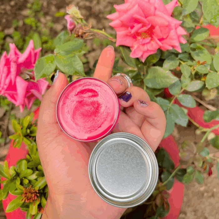 A hand with purple glitter nail art holding an open tin of Vaseline Lip Therapy Rosy Lips balm, showing the smooth pink product inside, with blooming pink roses and green leaves in the background.