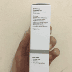 Hand holding the side view of The Ordinary Azelaic Acid Suspension 10% box, showing directions for use and visibly targeted concerns: uneven tone, dullness, and textural irregularities. White minimalist packaging against a beige wall background.