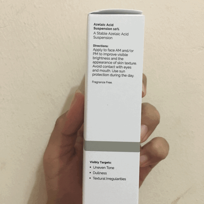 Hand holding the side view of The Ordinary Azelaic Acid Suspension 10% box, showing directions for use and visibly targeted concerns: uneven tone, dullness, and textural irregularities. White minimalist packaging against a beige wall background.