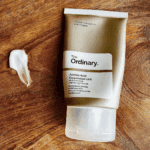 The Ordinary Azelaic Acid Suspension 10% in a grey squeeze tube placed on a wooden surface, with a small swatch of the brightening cream shown beside it to highlight texture.