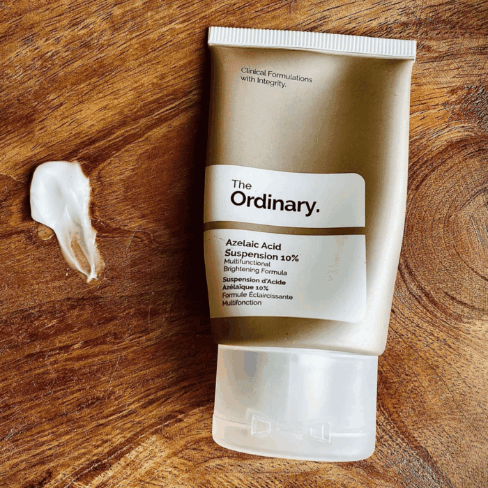 The Ordinary Azelaic Acid Suspension 10% in a grey squeeze tube placed on a wooden surface, with a small swatch of the brightening cream shown beside it to highlight texture.