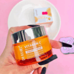 A hand with long black nails holding Dr Rashel Vitamin C Brightening & Anti-Aging Night Cream jar (orange with silver lid) against a pink background, with the cream’s box blurred in the background on a white plate.