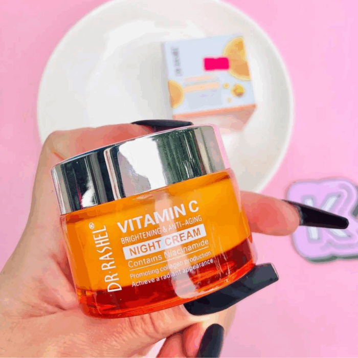 A hand with long black nails holding Dr Rashel Vitamin C Brightening & Anti-Aging Night Cream jar (orange with silver lid) against a pink background, with the cream’s box blurred in the background on a white plate.