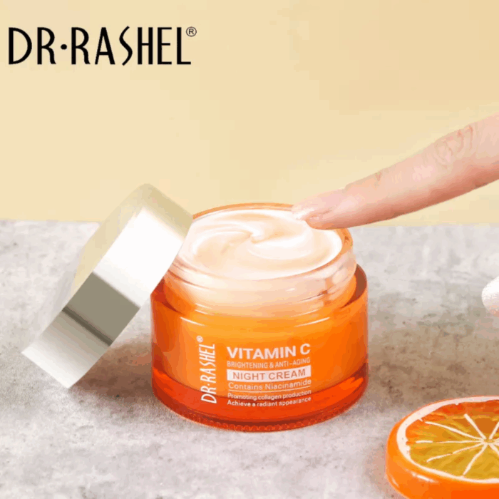An open jar of Dr Rashel Vitamin C Brightening & Anti-Aging Night Cream (orange with silver lid) on a gray surface, with a finger dipping into the soft cream. Background is beige with a dried orange slice on the side.
