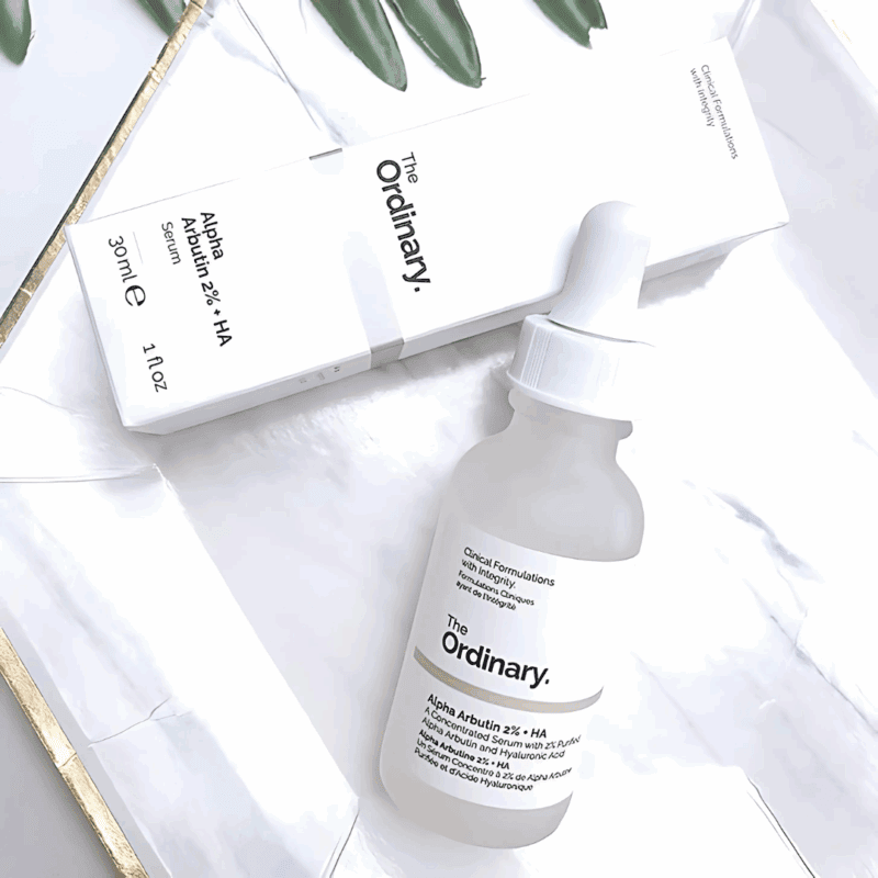 The Ordinary Alpha Arbutin 2% + HA Serum in a frosted glass dropper bottle placed on a white marble tray, with its minimalist white box packaging in the background and green leaves for a fresh look.