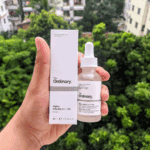 A hand holding The Ordinary Alpha Arbutin 2% + HA Serum in a frosted glass dropper bottle alongside its white box packaging, with a green leafy outdoor background.