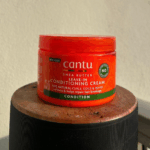 Cantu Shea Butter Leave-In Conditioning Cream jar placed on a wooden surface, designed to soften curls, coils, and waves while repairing hair breakage.