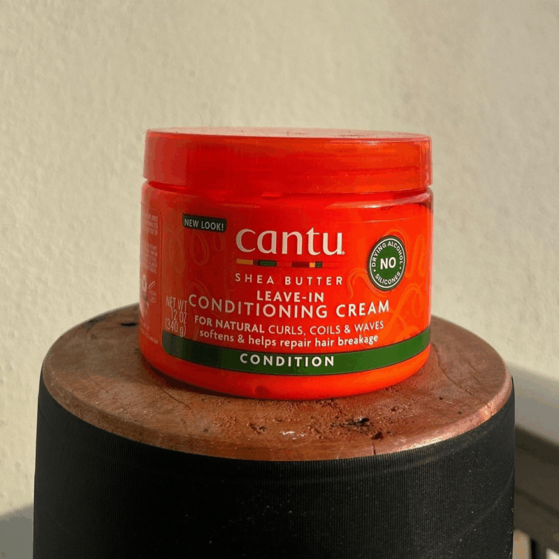 Cantu Shea Butter Leave-In Conditioning Cream jar placed on a wooden surface, designed to soften curls, coils, and waves while repairing hair breakage.
