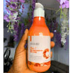 Hand holding Dr Rashel Vitamin C Brightening & Nourishing Body Lotion 300ml bottle with floral background.