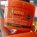 Hand holding Cantu Shea Butter Leave-In Conditioning Cream jar, formulated to soften, condition, and repair breakage in curly, coily, and wavy hair.