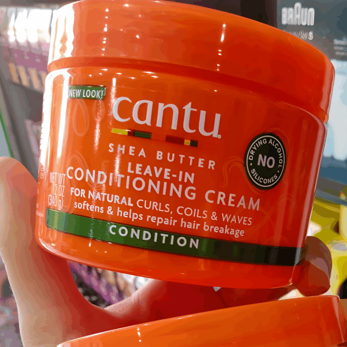Hand holding Cantu Shea Butter Leave-In Conditioning Cream jar, formulated to soften, condition, and repair breakage in curly, coily, and wavy hair.