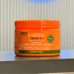 Cantu Shea Butter Leave-In Conditioning Cream jar placed on a table, designed to condition, soften, and repair breakage for natural curls, coils, and waves.
