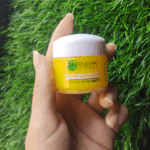 Hand holding Garnier Bright Complete Vitamin C Sleeping Mask Night Cream jar against a green grass background.