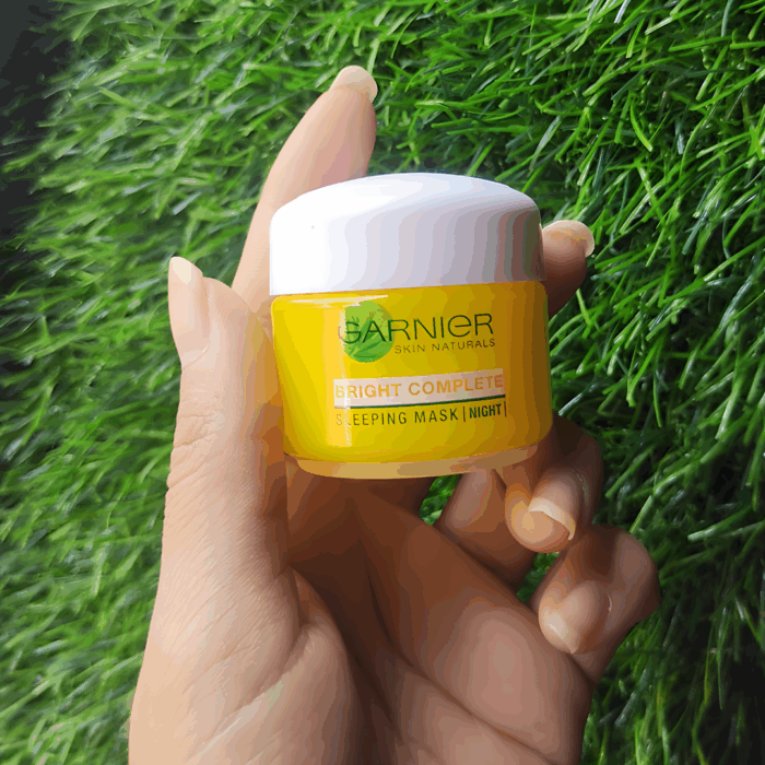 Hand holding Garnier Bright Complete Vitamin C Sleeping Mask Night Cream jar against a green grass background.