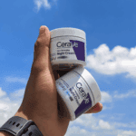 A hand holding two jars of CeraVe Skin Renewing Night Cream against a bright blue sky with scattered clouds.
