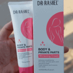 Close-up of a hand holding the Dr Rashel Body & Private Parts Whitening Cream tube next to its box, with pink and white packaging featuring a feminine silhouette design.