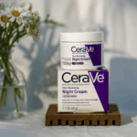 A jar of CeraVe Skin Renewing Night Cream placed on top of its box, displayed on a wooden stand with soft natural lighting and flowers blurred in the background.