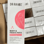 A close-up of the Dr Rashel Body & Private Parts Whitening Cream box held above a carton filled with multiple identical boxed products.