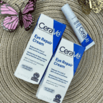 CeraVe Eye Repair Cream boxes and tube displayed on a textured surface with a butterfly decoration, highlighting its benefits for reducing puffiness, dark circles, and restoring moisture around the eyes.