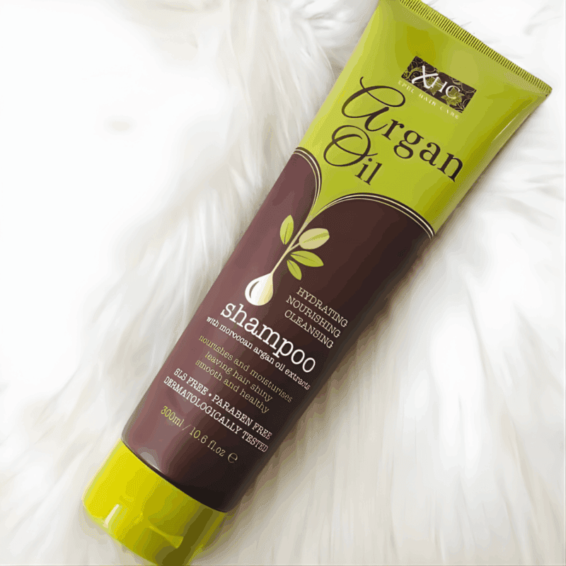 XHC Argan Oil Shampoo tube placed on a white fur background, featuring green and brown packaging. The label highlights Moroccan argan oil extract for hydrating, nourishing, and cleansing hair, and mentions it is SLS-free and paraben-free.