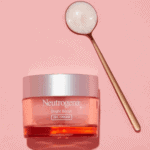 Neutrogena Bright Boost Gel Cream in a pink jar with a silver lid placed on a pastel pink background beside a gold spoon containing the cream’s pearly texture, symbolizing brightness and glow.