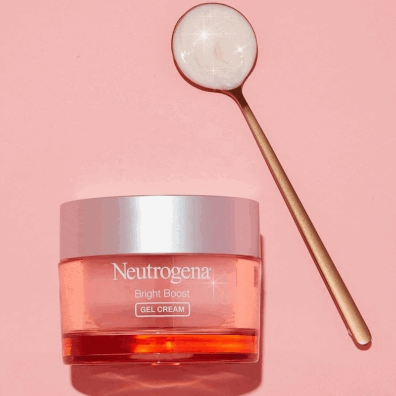Neutrogena Bright Boost Gel Cream in a pink jar with a silver lid placed on a pastel pink background beside a gold spoon containing the cream’s pearly texture, symbolizing brightness and glow.
